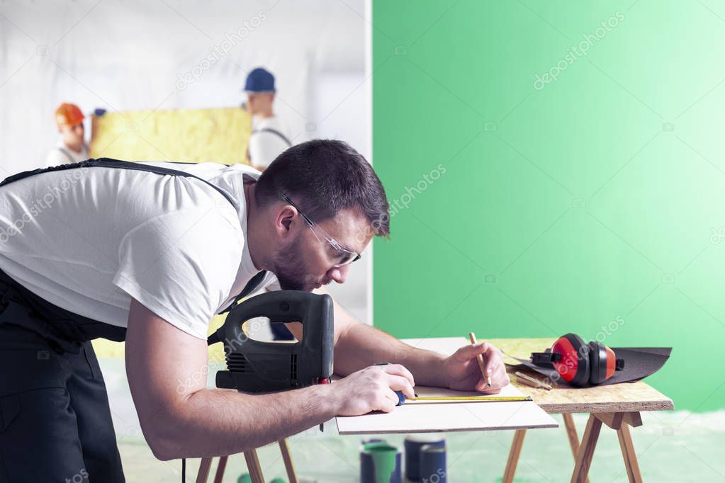 Construction worker measuring a wooden plank using a tape measure. Place your logo on a green wall