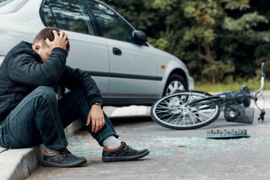 Kafasını tutarak ve bir araba ve bisiklet kazasında yanındaki kaldırımda oturan endişeli motorcu