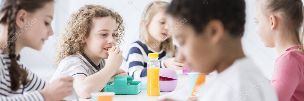 Panorama of school kids eating lunch together during a break, at a school canteen. Focus on a boy with curly hair eating a sandwich