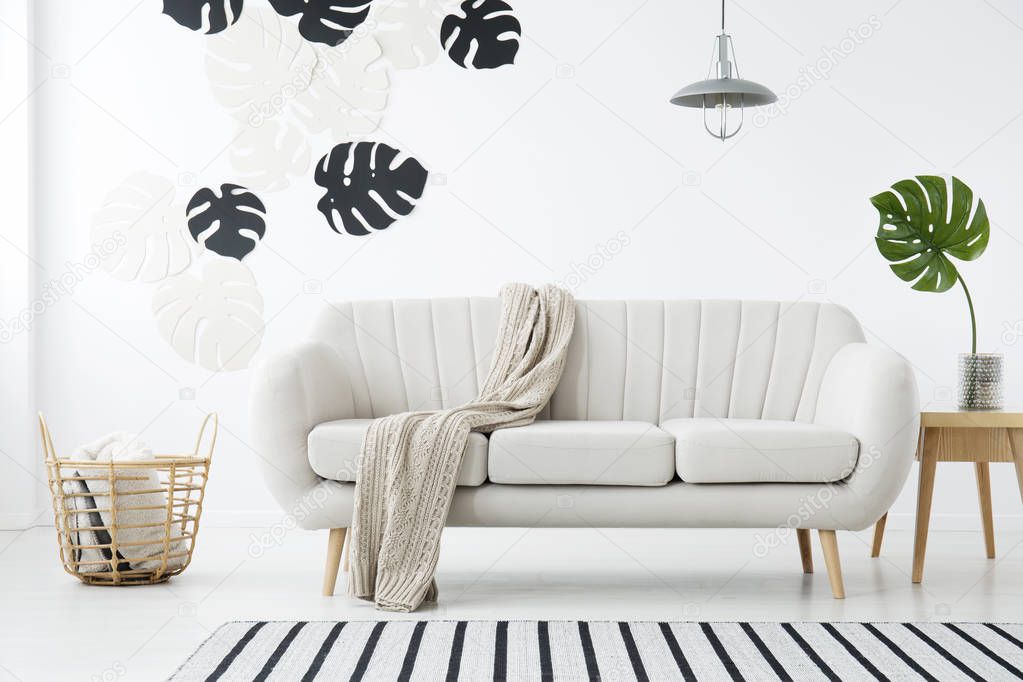 Blanket on sofa next to basket in white living room interior with monstera leaf on table. Real photo