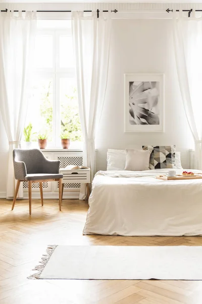 ウィンドウの敷物と寝室のインテリアのポスターの下に白いベッドの横にある灰色のアームチェア 実際の写真 — ストック写真