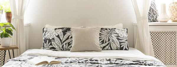 Panorama close-up of a large bed with beige and black pillows with floral pattern in a bright bedroom interior with white walls and decor