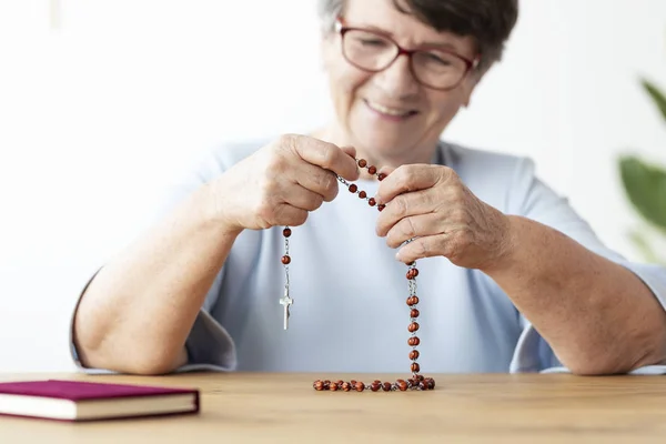 微笑的宗教老年妇女祈祷与念珠在餐桌上与圣经 专注于手 — 图库照片