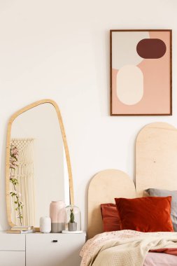 Mirror on white cabinet next to bed with red cushions under poster in pastel bedroom interior. Real photo clipart