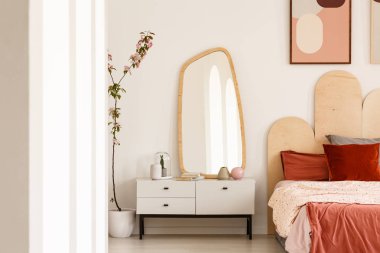 Plant next to white dressing table with mirror in red bedroom interior with poster above bed. Real photo clipart