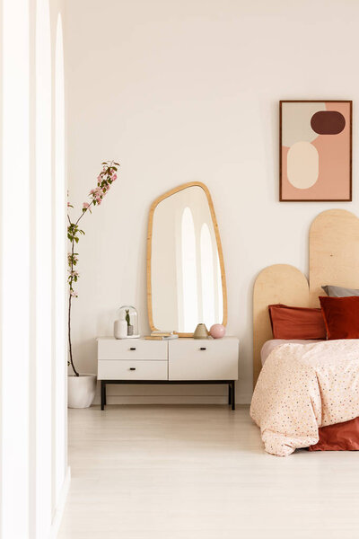 Mirror on white cabinet next to bed with headboard under poster in bright bedroom interior. Real photo