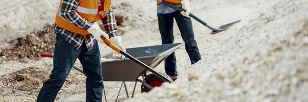 反射安全ベストと砂を掘りでサイトの建設労働者のパノラマは 手押し車にシャベルします — ストック写真