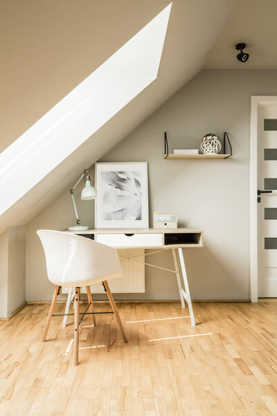 White chair standing by the wooden desk with pastel lamp and simple poster in real photo of bright attic room interior