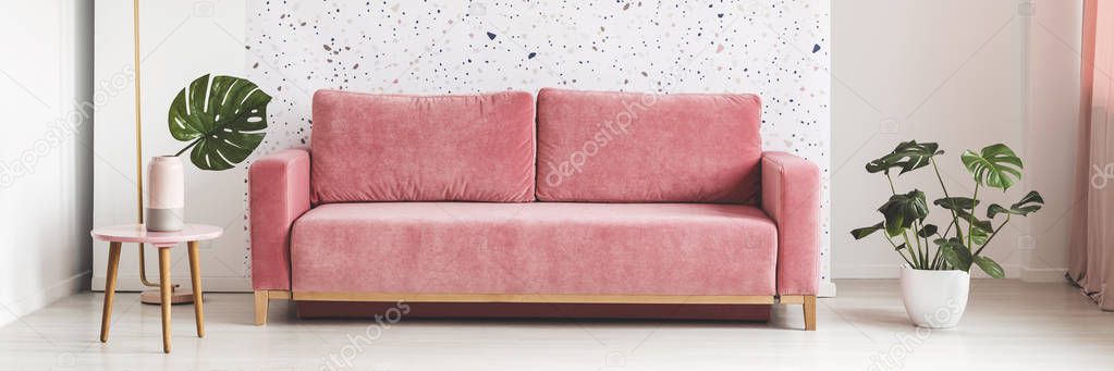 Pink velvet sofa in real photo of bright living room interior with lastrico pattern wall and Monstera Deliciosa leaf