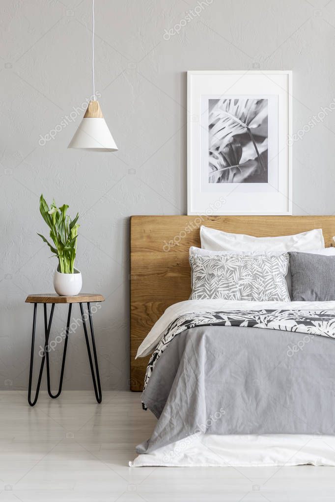 Plant on table next to wooden bed in grey bedroom interior with lamp and poster. Real photo