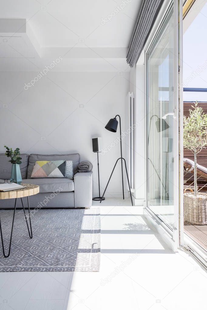 Real photo of white living room interior with grey couch, black lamp, two posters and glass door to the terrace