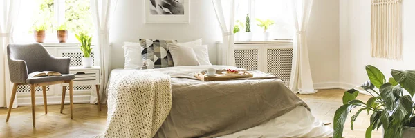 木製トレイ朝食とニットのブランケット 緑の植物と本と灰色のアームチェア キングサイズのベッドと明るい寝室のインテリアの本物の写真 — ストック写真