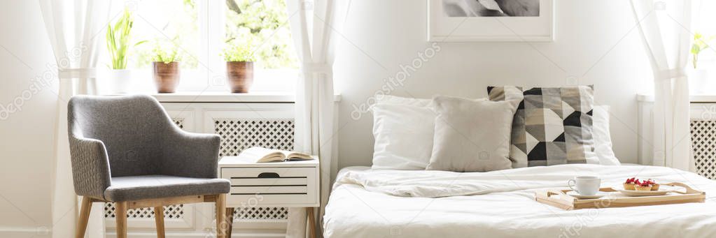 Grey armchair standing by the bedside table with open book in the real photo of white bedroom interior with double bed with cushions and white sheets and plants on windowsill