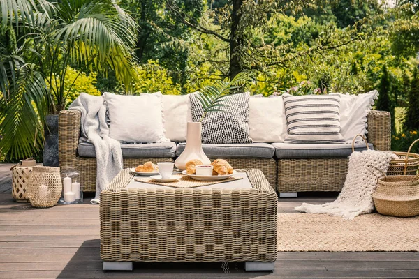 Rattan table and pillows on couch standing on a patio in the garden during summer. Real photo