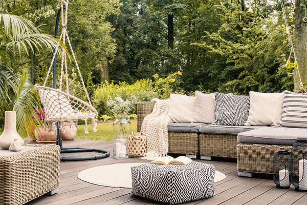 Book on a black and white pouf in the middle of a bright terrace with a rathan corner sofa, hanging chair and round rug. Настоящее фото
