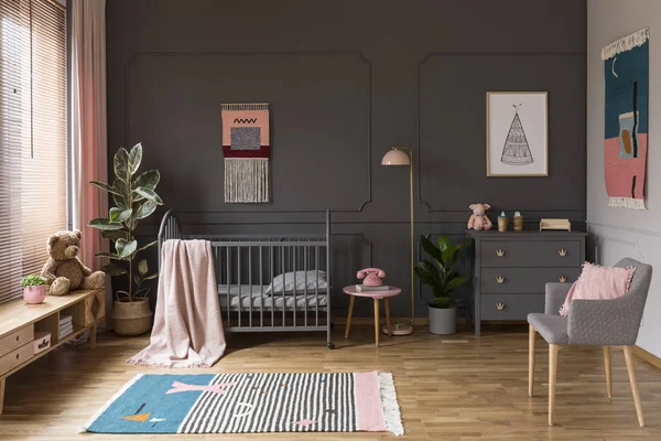 真正的照片 一个灰色的婴儿床站在粉红色的凳子旁边 一盏灯和橱柜在灰色婴儿室内部也与扶手椅 地毯和海报 — 图库照片