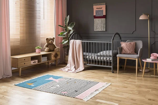 婴儿婴儿床的真实照片站在一个低矮的橱柜和扶手椅 儿童房内的木地板和灰色墙壁与成型的灯和凳子之间 — 图库照片