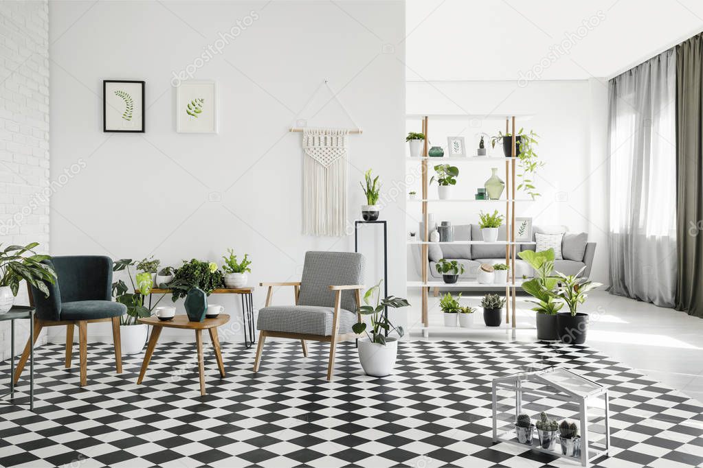 Wooden table between armchairs on checkered floor in white living room interior with plants. Real photo