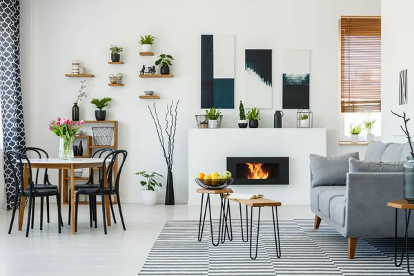 Chairs at table with flowers in white apartment interior with grey sofa next to fireplace. Real photo