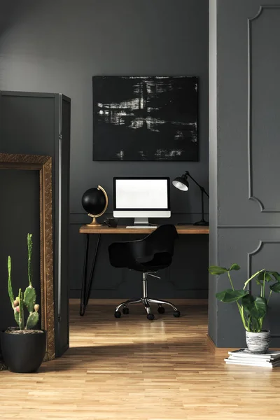 Dark living room interior with molding on walls in real photo with green plants, modern painting and home office desk with empty screen computer