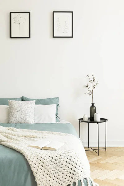 Posters above bed with pillows and knit blanket in bright bedroom interior with table. Real photo