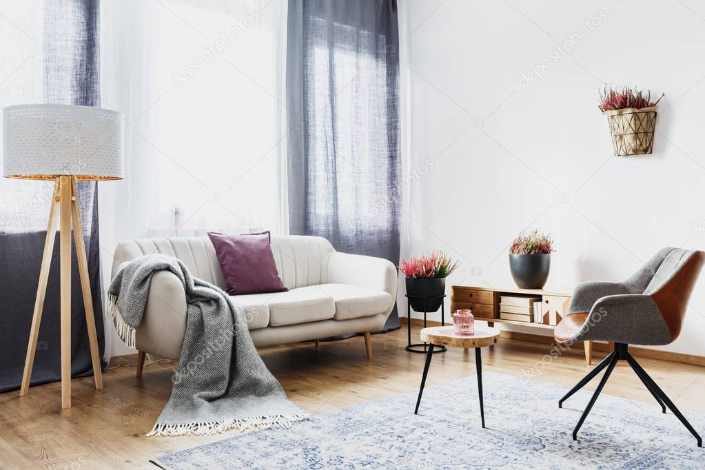Blanket and pillow on a sofa next to a wooden lamp in cozy flat interior with heathers and grey armchair