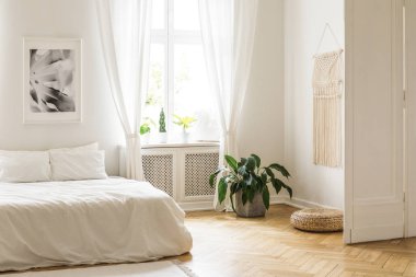 Beige macrame and a poster on the white walls of a bright, minimalist bedroom interior with herringbone hardwood floor and plants