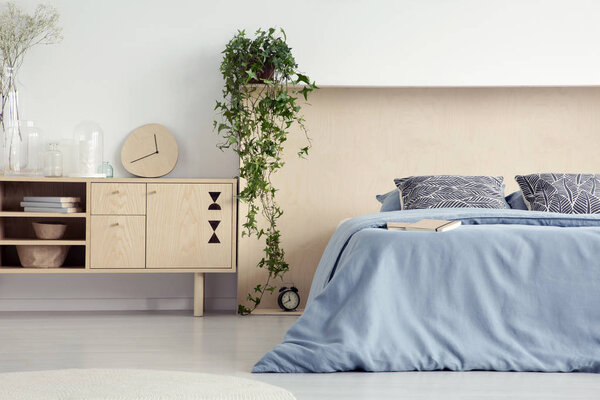 Clock on wooden cupboard next to to blue bed with patterned pillows in bedroom interior. Real photo