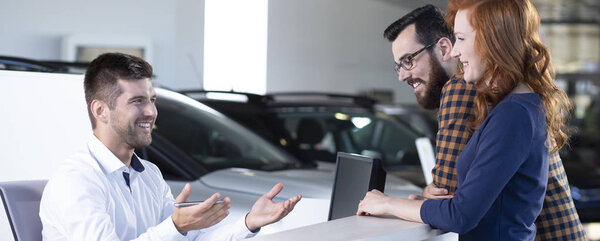 Panorama of smiling dealer offering new car to happy buyers in a salon
