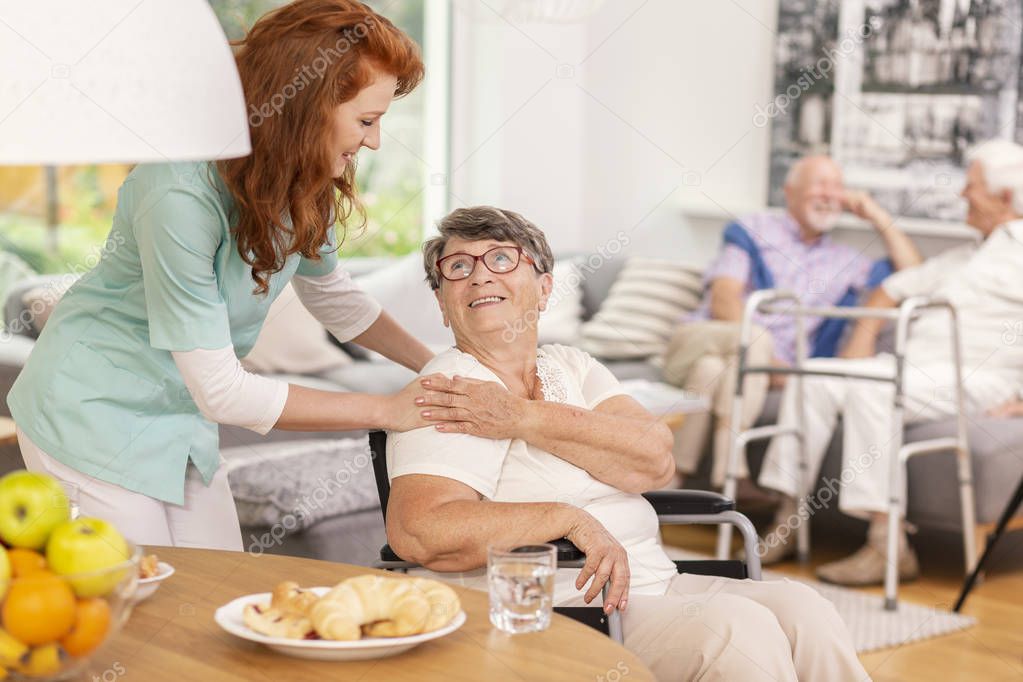 Friendly nurse supporting smiling senior woman in nursing house. Blurred background