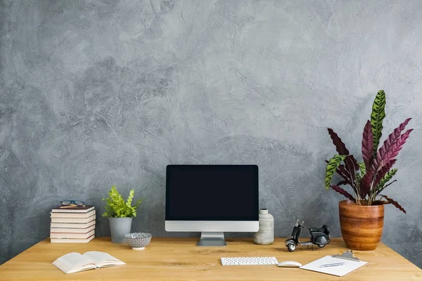 Plant, computer and books on a wooden desk set on a grey wall. Product placement