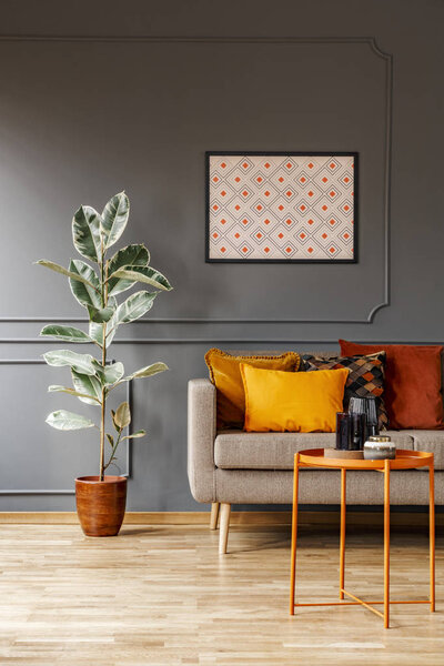 Real photo of poster with geometric pattern hanging on the wall with wainscoting in dark living room interior with fresh plant, sofa with cushions and metal end table with decorations