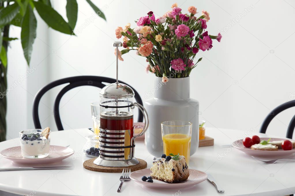 Real photo of white dining table with fresh flowers in ceramic vase, pink plates with cake and juice in glass