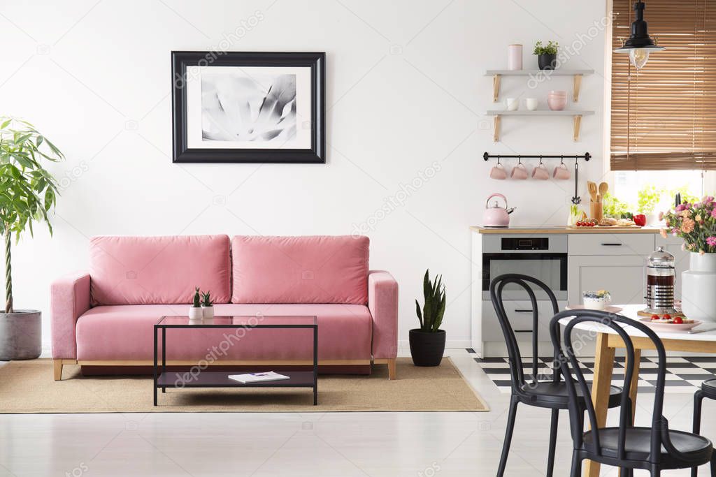 Poster above pink couch in white apartment interior with black chairs at table and kitchenette. Real photo