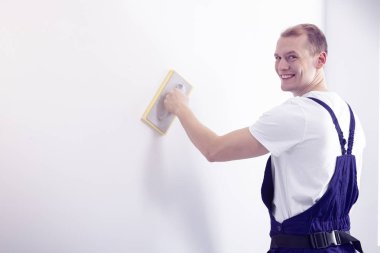 Genç, mutlu aşağı beyaz bir duvar bir zımpara aracıyla düzgünleştirme kameraya bakarak ressam iş giysisi poz içinde duvar