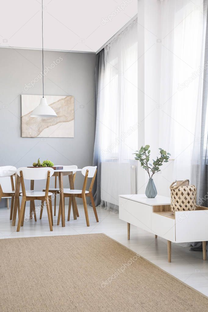 White chairs at dining table in scandi flat interior with cupboard, painting and carpet. Real photo