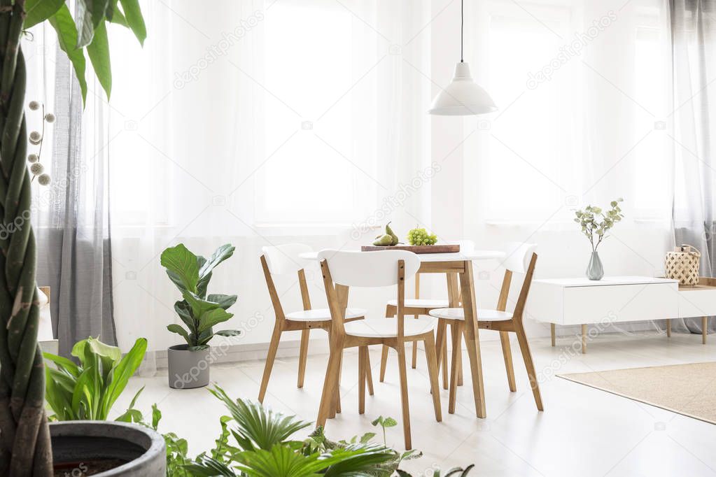 Real photo of a bright, scandi dining room with a white lamp above a round table with wooden chairs, next to the window