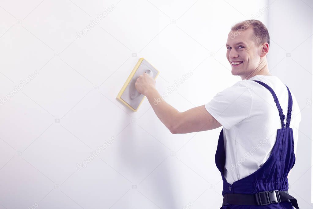 Young, happy wall painter in workwear posing, looking at the camera while smoothing down a white wall with a sandpaper tool