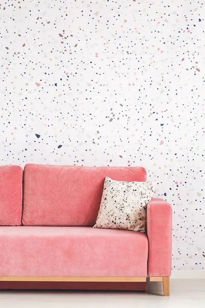 Pillow on pink sofa against patterned wallpaper in bright living room interior. Real photo
