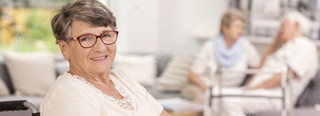 Panorama of happy elderly woman in the nursing house. Blurred seniors in the background