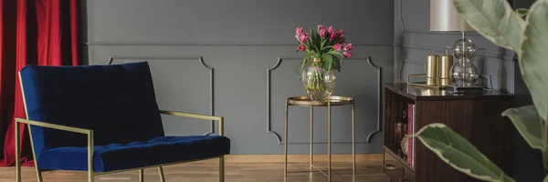 A luxurious, modern, navy blue armchair and a bouquet of pink tulips on a golden table in an elegant living room interior. Real photo.