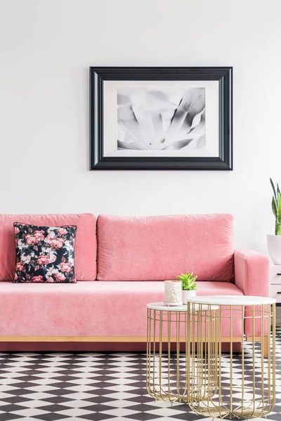 Velvet couch with a floral pillow, poster on the wall and metal coffee tables in a living room interior with checkered floor. Real photo