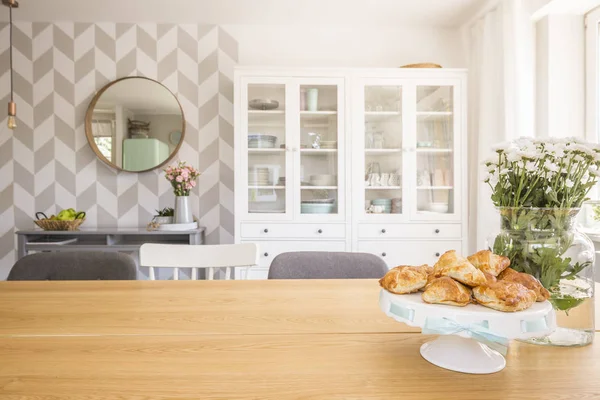 Croissants on wooden dining table in white apartment interior with flowers and mirror. Real photo