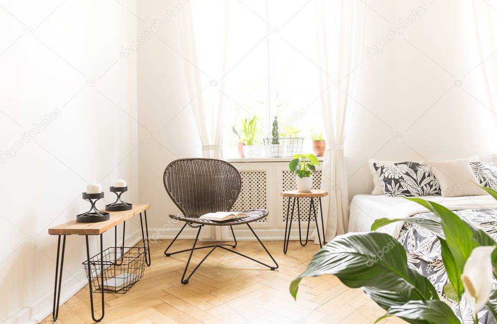 Bright light shining through a window into a modern bedroom interior with a rattan and metal chair, bedside stand with candles and a bed. Copy space on white wall. Real photo.