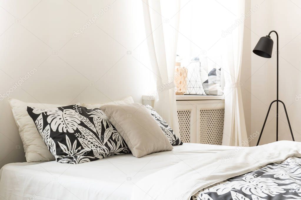 Close-up of pillows and leaf motif cover on a bed in a bright bedroom interior. Black metal lamp next to the bed. Real photo.