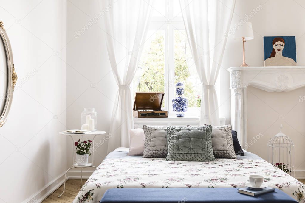 Window with drapes in real photo of white bedroom interior with king-size bed with pillows, retro gramophone on windowsill and bedside table with fresh flowers and candles