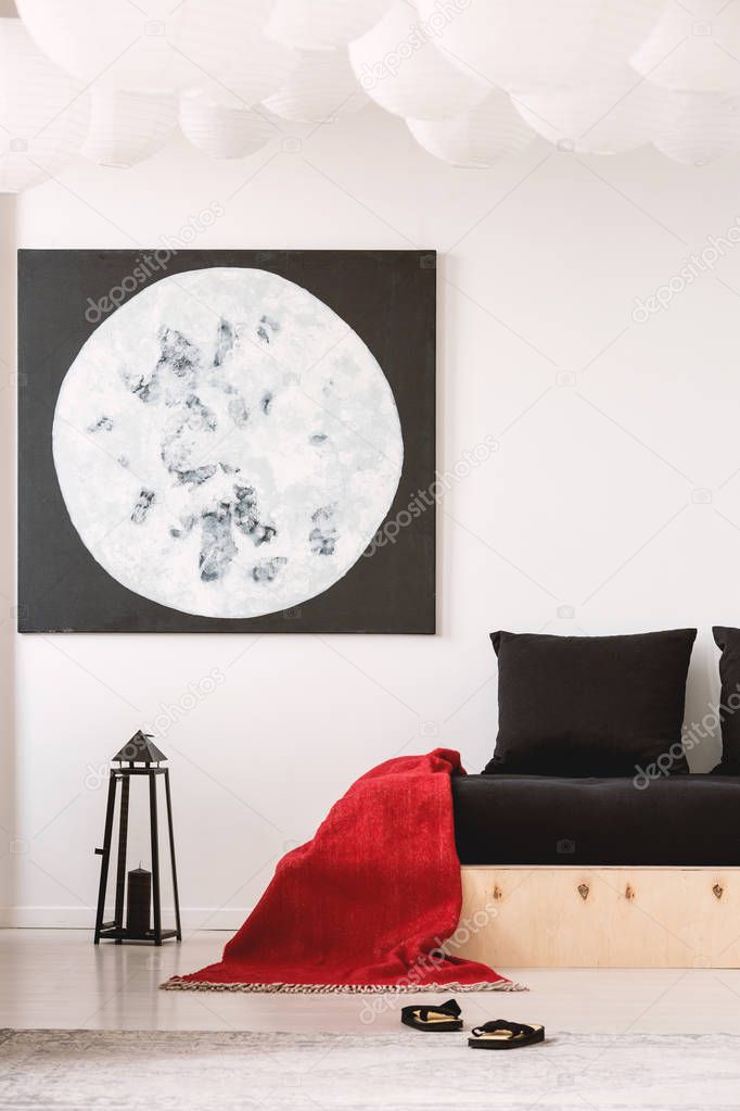 Red blanket on black wooden bed in white bedroom interior with moon poster above lantern. Real photo