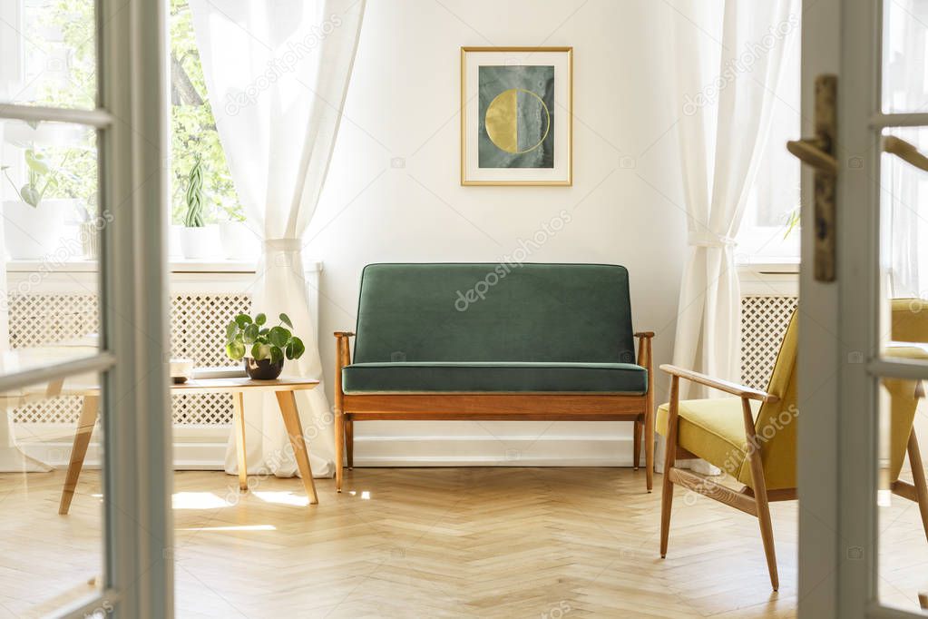 Real photo of a vintage living room interior with a green sofa, poster, coffee table, armchair and wooden floor. View through a door
