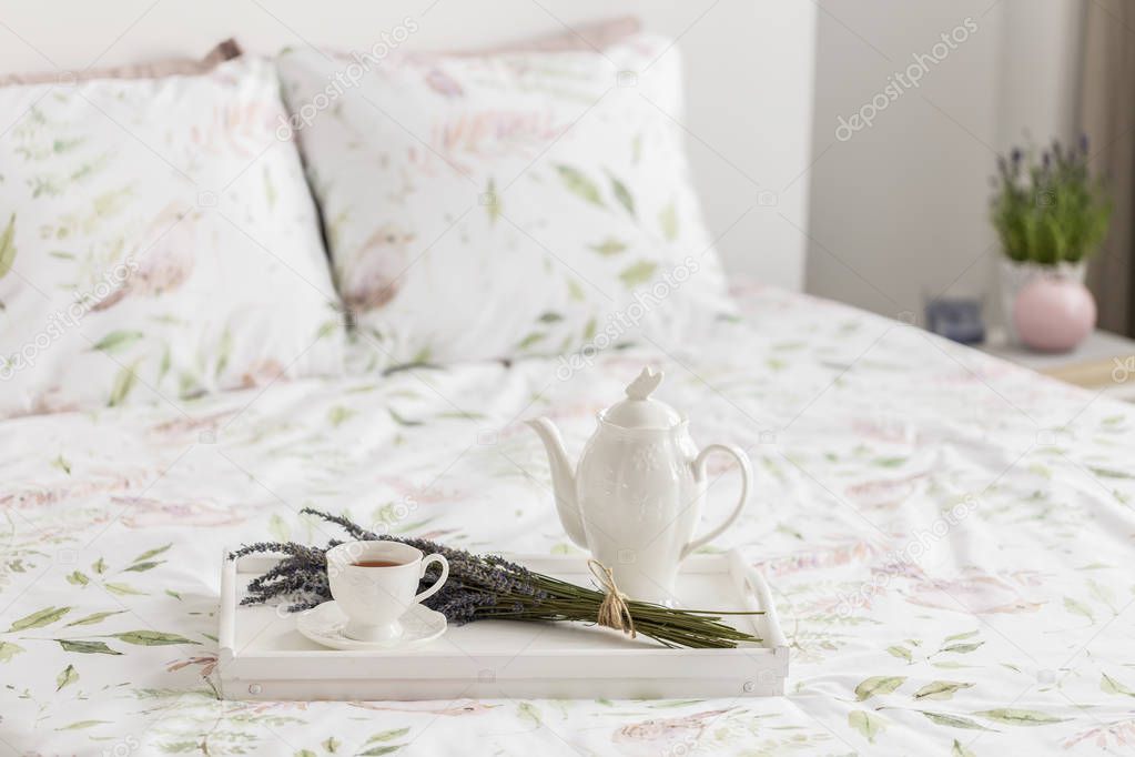 Real photo with close-up of breakfast tray with tea cup, jug and lavender placed on double bed with floral bedclothes. Blurred background