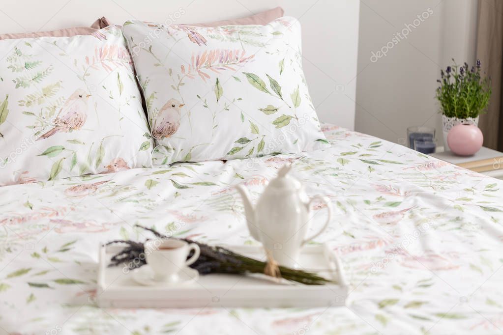 Close-up of a blurred tray with a pot, cup and bouquet of flowers on a double bed in a bedroom interior